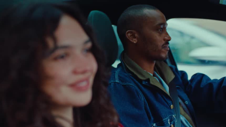 Young adult woman sitting on the passenger seat of a car next to a young adult black man driving and occasionally looking at her while both smile during day time