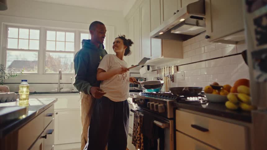 A man wearing a green hoodie and a woman wearing a white t shirt prepare food together at a stove in a bright kitchen during the day