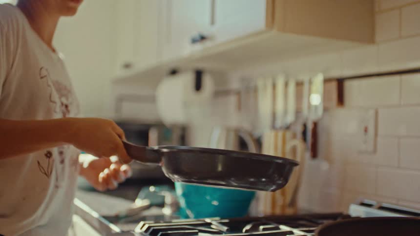 A woman wearing a white t shirt tosses a pancake in a frying pan while standing in a kitchen