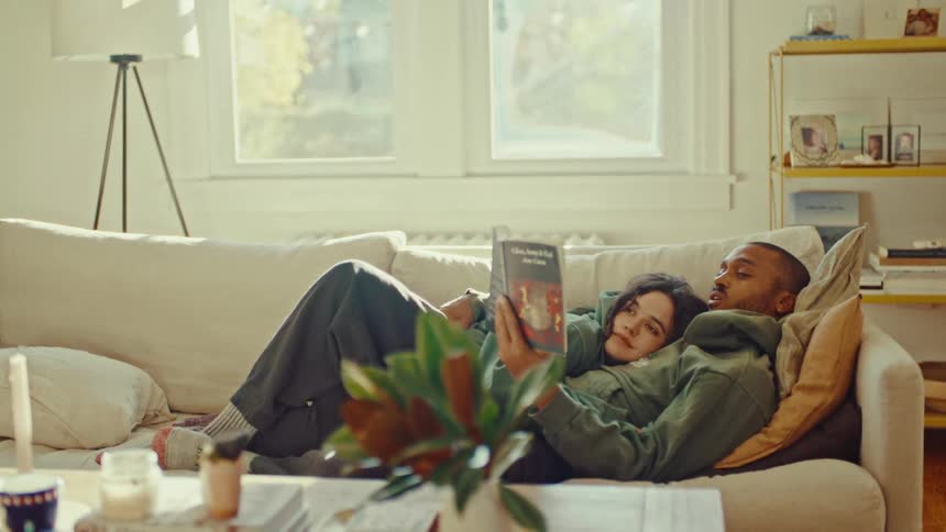 A man wearing a green hoodie and a white cap lies on a couch while a woman rests against him as they look at a book together in a bright living room
