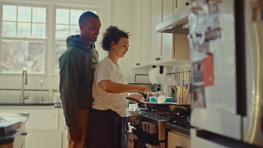 A couple stands closely together in a sunlit kitchen smiling and cooking at the stove