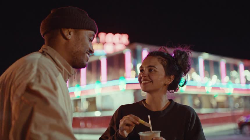 A happy woman stands outside a well lit restaurant feeding a man fries dipped in a shake