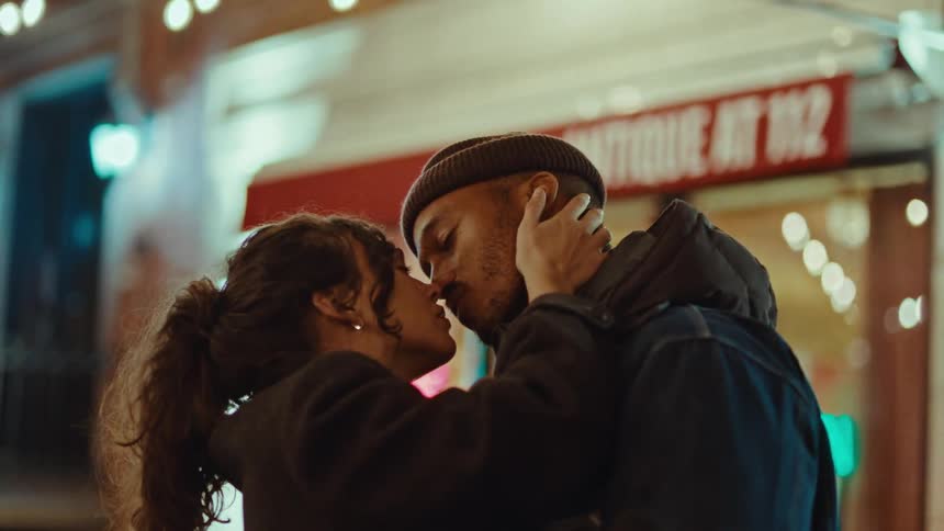 A couple shares an intimate kiss on a city street at night with the woman gently cradling the mans face in her handsthe warm blurred lights of a storefront labeled antique at 112 create a romantic and cinematic atmosphere behind them