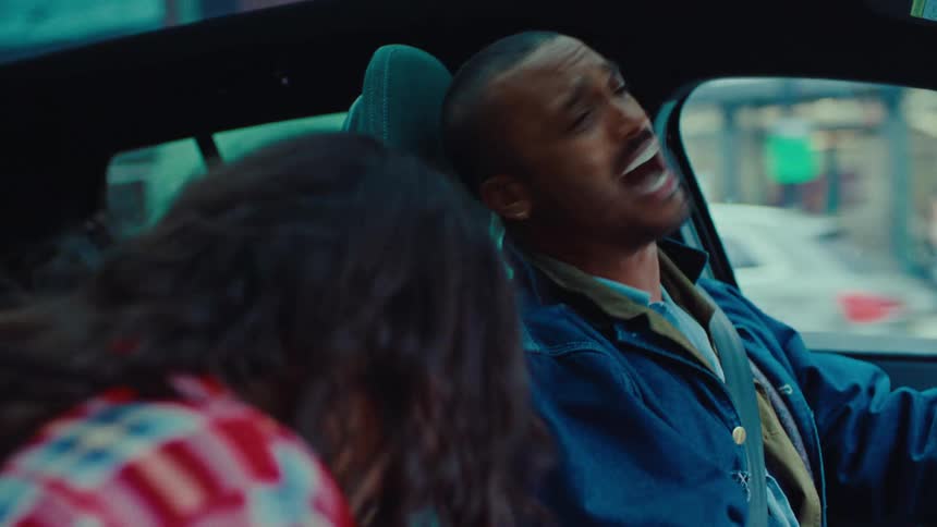 A man in a denim jacket sits in the drivers seat of a car singing with great enthusiasm and expressive facial gesturesnext to him a woman in a red plaid shirt watches and reacts as they share a lively moment while driving