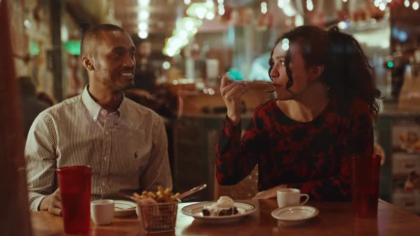 A man and woman sit at a wooden table in a dimly lit cafe talking and smiling as she eats a brownie sundae