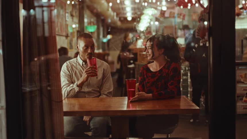 A man and a woman sit at a wooden table in a dimly lit restaurant sharing a conversation and drinks from red cupsthe man takes a sip of his drink while the woman gestures with her hand both appearing relaxed in the warm casual atmospher