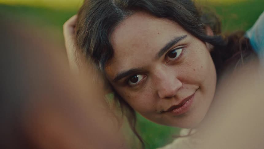 A young woman lies in a grassy field looking tenderly at someone just out of frameshe alternates between a soft lingering gaze and a gentle smile as she tucks a lock of hair behind her ear