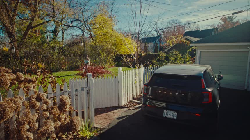 A black volvo suv sits parked in a suburban driveway while two people are playing on a grassy backyard behind a white picket fence on a clear autumn day