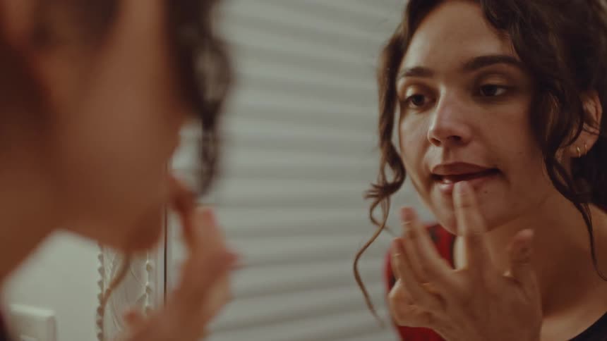 A young woman with dark curly hair looks into a white framed mirror while carefully applying lip liner and blending it with her finger