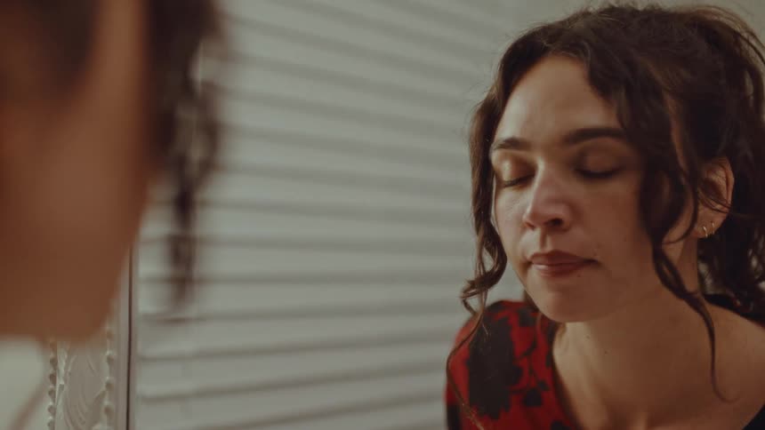 A woman wearing a red floral shirt looks into a mirror while adjusting her hair in a room with white blinds