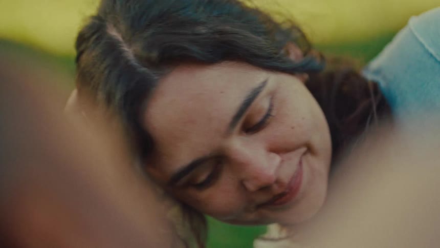 A woman with wavy brown hair smiles while lying on green grass in a park during the day