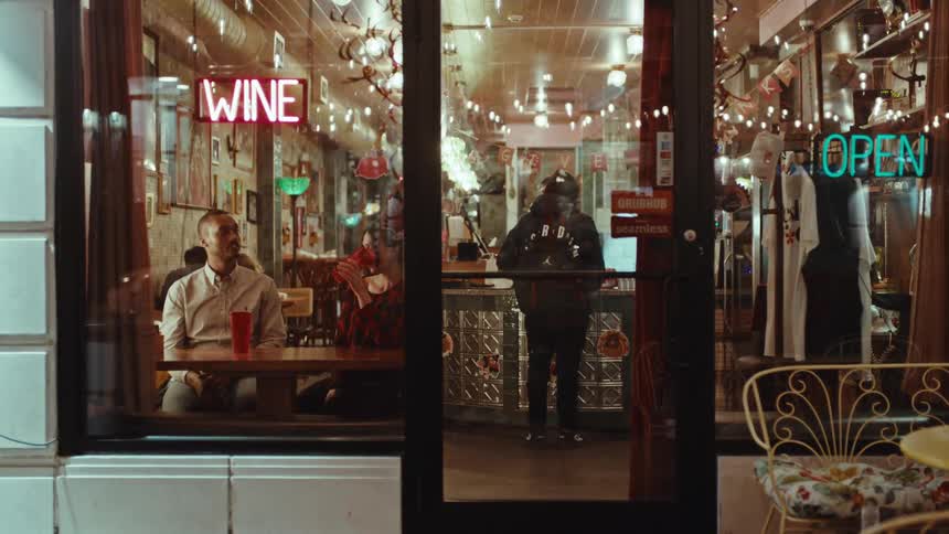Young adult man and woman sitting down and talking while drinking from tall cups in a bar with other customers and servers on the background seen through a window