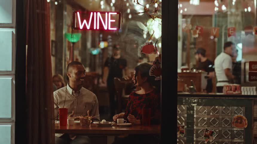 A smiling happy couple sit inside a cafe eating and drinking while several workers are around in the restaurant working