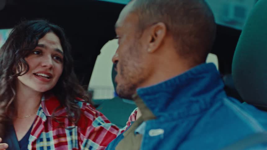 A young woman and a black man are traveling inside a vehicle singing smiling and gesturing