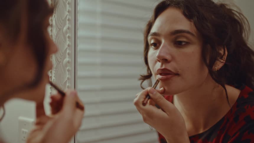 A woman with dark curly hair looks into a mirror while carefully applying a lip pencil to her lips as she stands in front of a window with white blinds and a white wall switch