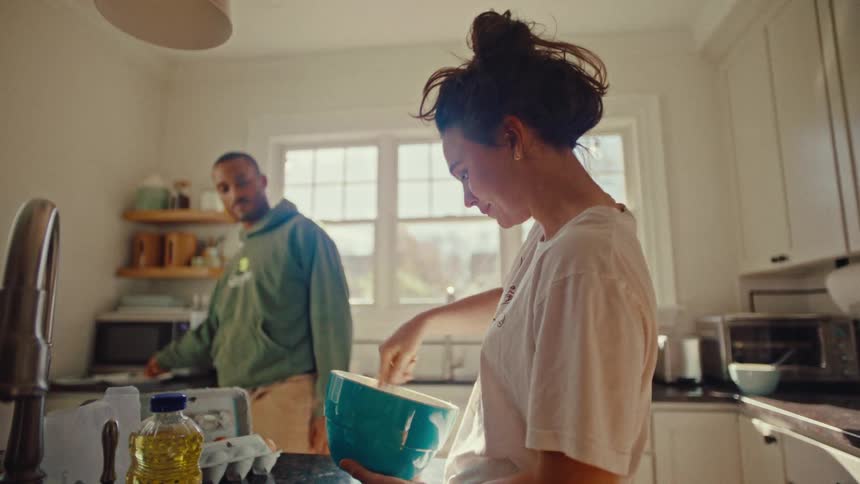 A woman with her hair in a bun stirs a mixture in a blue bowl while a man in a green hoodie stands in the background of a bright kitchen