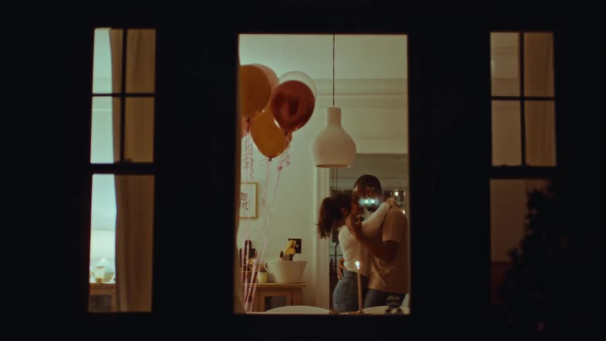 Young adult man and young adult woman dancing and hugging while smiling in a living room with balloons and candles seen through a window during night time