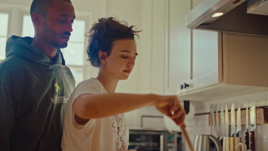 A man wearing a green hoodie stands behind a woman wearing a white t shirt as she stirs a pot with a wooden spoon while they cook together in a bright kitchen