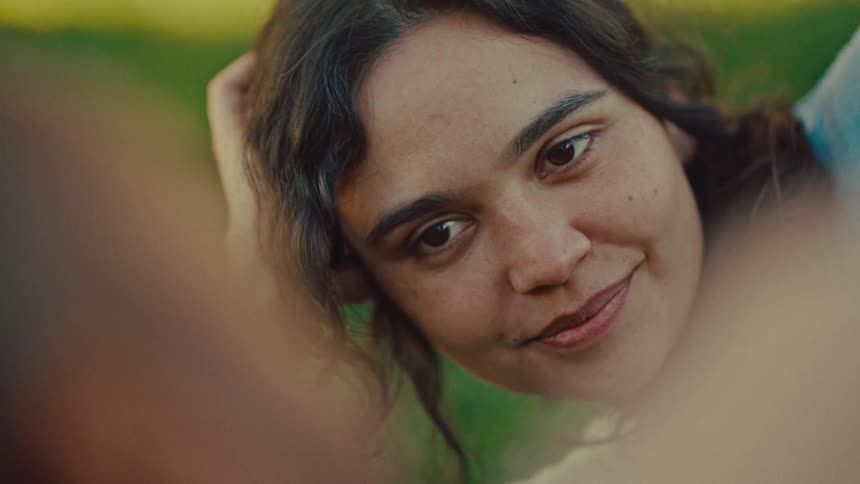 A woman with wavy brown hair looks down and smiles while lying on green grass in a park during the day