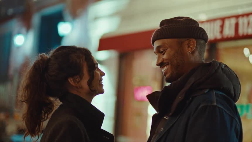 Young adult couple talking and laughing before starting to kiss slowly until moving to a more passionate slow kiss outdoors during night time