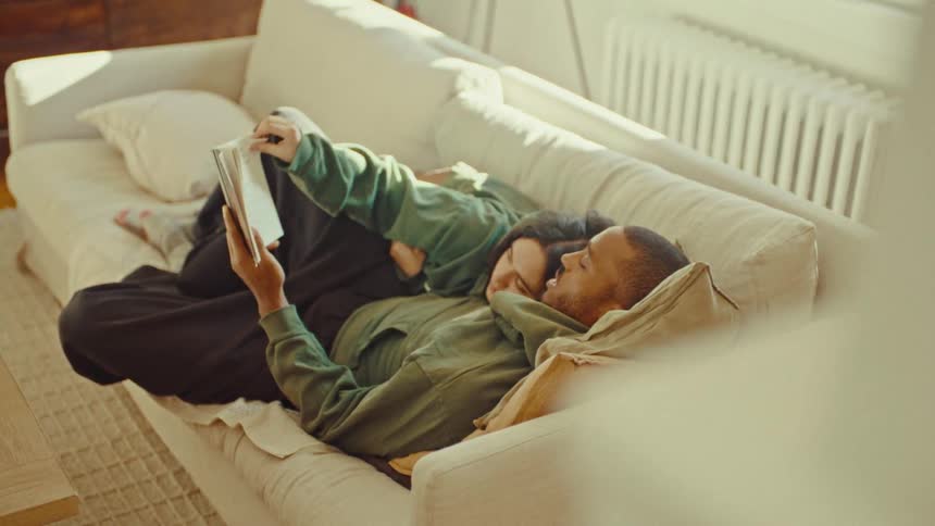 Man lying on a couch reading a book while a woman rests beside him