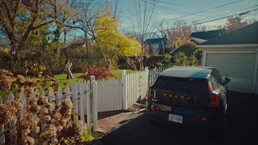 A black electric car is parked in a driveway next to a white picket fence while a couple play on a green lawn in a suburban backyard on a sunny day