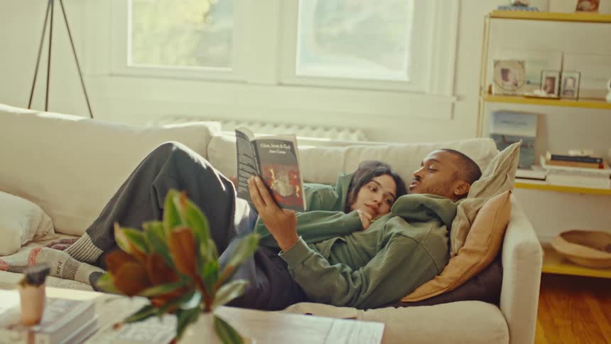 Young adult black man lying on a couch and reading aloud with a young adult woman resting on his chest in a living room indoors during day time