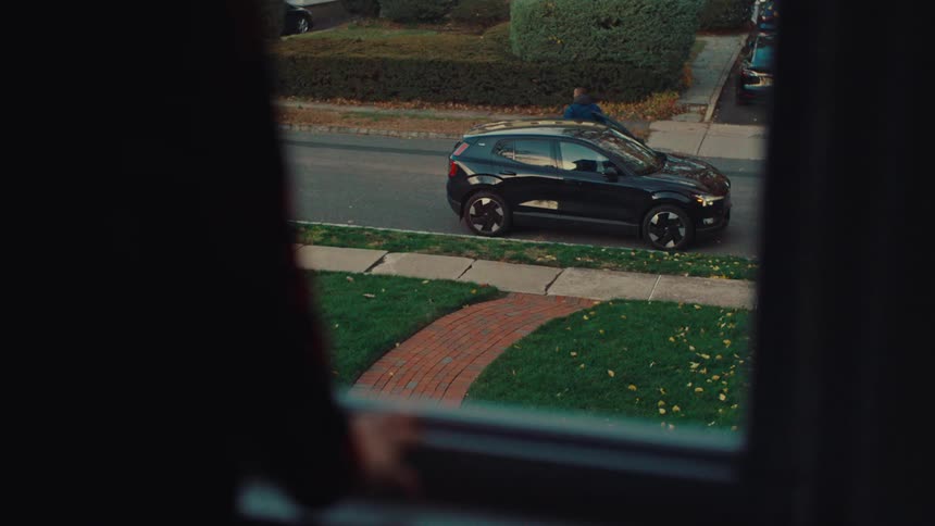 A person from the shoulders down is seen standing at a window watching a male park and get out of the suv and walk to the house