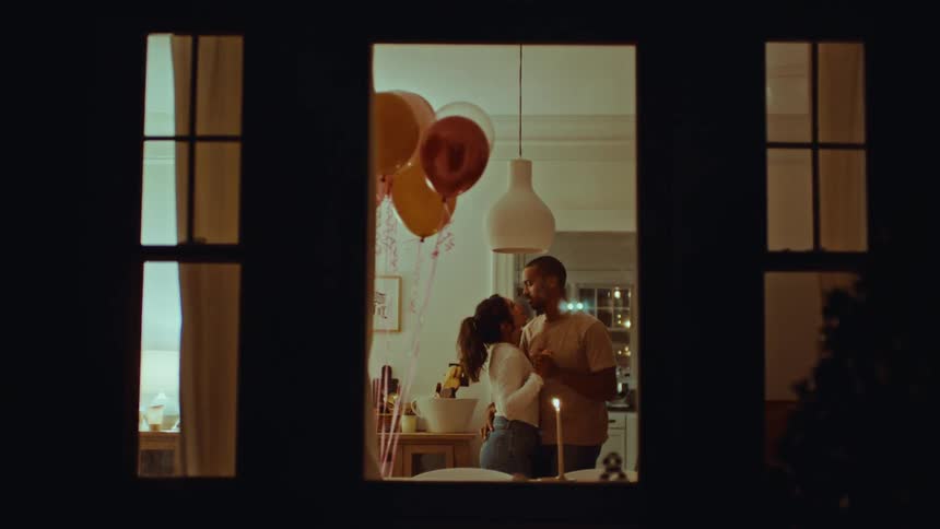 A couple dances slowly in their kitchen framed by a window and surrounded by festive balloons they share a tender kiss and smile at each other during their intimate celebration