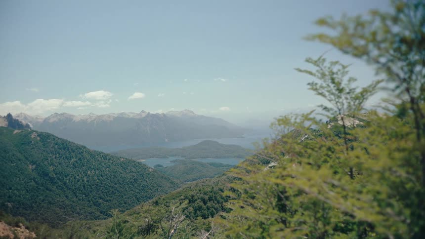 A wide panoramic view reveals lush green forested mountains sloping down toward a large blue lakein the far distance a range of snowcapped mountain peaks sits beneath a clear pale sky