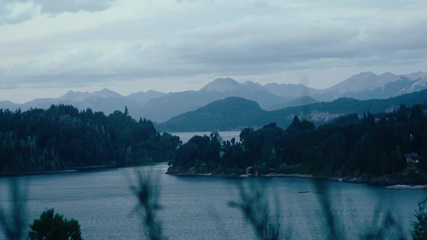 Dense green forests and rolling hills surround a wide calm lake under a cloudy sky jagged mountain peaks line the distant horizon as soft light filters through the overcast atmosphere