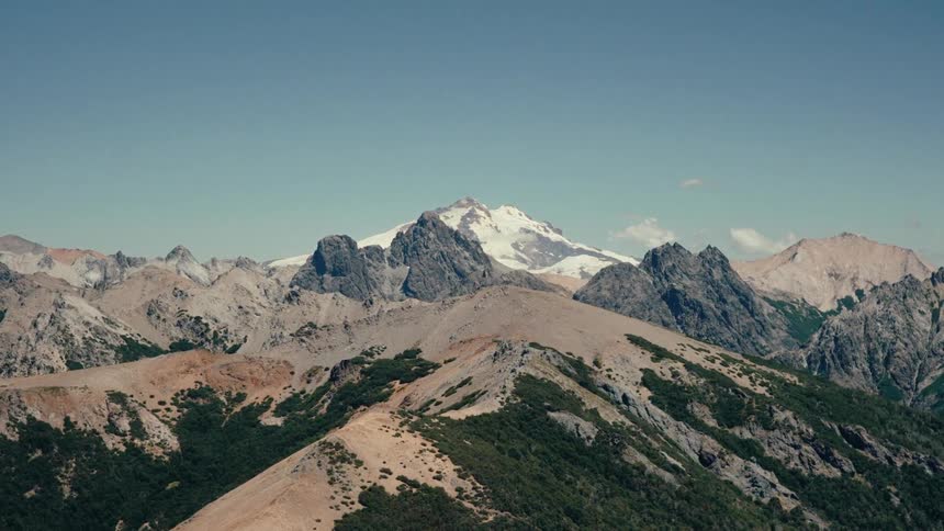 A rugged mountain range under a blue sky where a massive peak covered in white snow and ice towers over the surrounding sharp grey ridges and brown slopes dotted with small green bushes