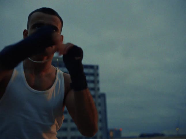 A man wearing a white tank top and black hand wraps practices boxing on a rooftop during twilight with large buildings and city lights visible in the background