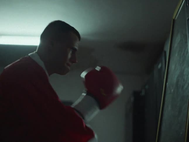 A man wearing a red and white robe with red boxing gloves shadow boxes in a dimly lit locker room under a fluorescent light