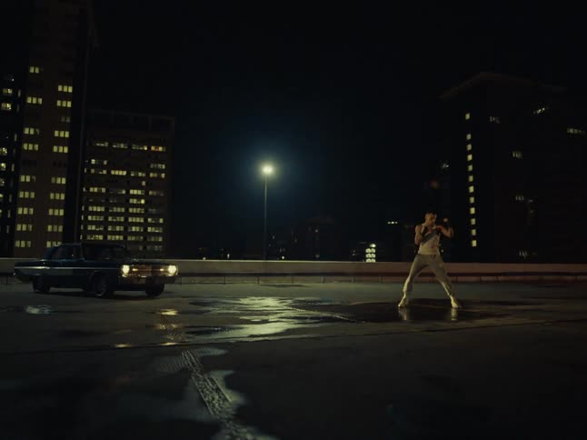 A man in a white tank top and grey pants shadow boxes on a wet rooftop at night while a dark car sits parked nearby against a city backdrop