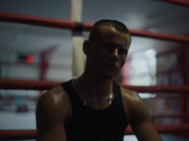 A weary boxer sits alone on the edge of the ring catching his breath in the dimly lit atmospheric gymthe camera moves into a tight closeup capturing the sweat on his skin and a look of intense quiet focus