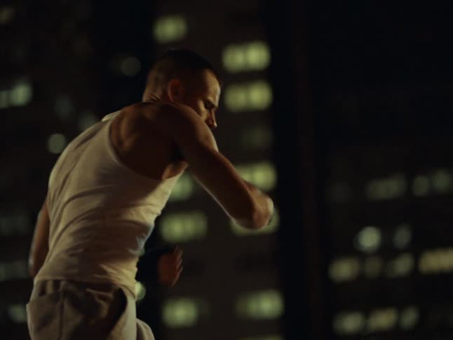 A man wearing a white tank top and grey pants shadow boxes on a wet rooftop at night while a dark car sits parked nearby against a city backdrop