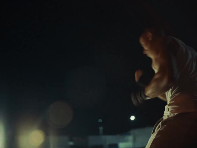 A man wearing a white tank top and gray trousers performs boxing movements and shadow boxes outdoors at night with distant city lights and a lens flare in the background