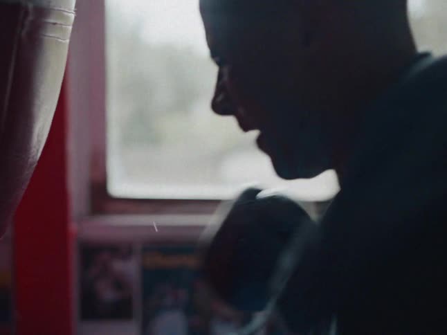 The video shows a closeup side profile of a person wearing a grey hoodie and boxing gloves they are moving their arms in a rhythmic training motion in front of a window