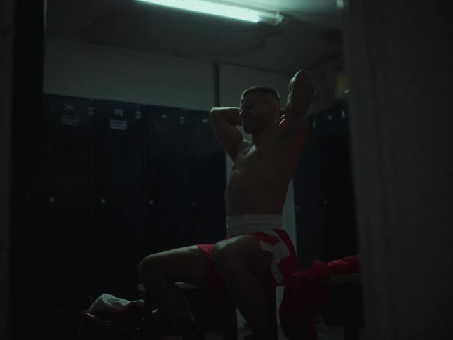 A male boxer sits on a bench in a dark locker room while stretching his arms and neck