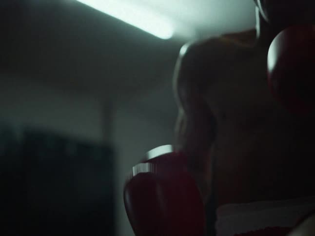 In a dimly lit gym a shirtless boxer wearing vibrant red gloves intensely shadowboxes throwing quick rhythmic punches toward the camera as a single overhead light casts dramatic shadows across his moving frame
