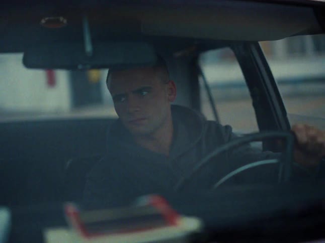 A man wearing a dark hoodie drives a car during the day while looking ahead and turning the steering wheel with a serious expression