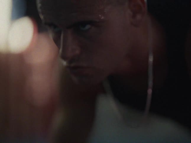 A sweaty man in a black tanktop performs pushups while staring intensely into the camera