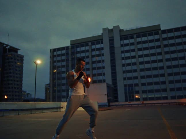A man wearing a white tank top and grey pants shadow boxes on a rooftop at night against a city backdrop