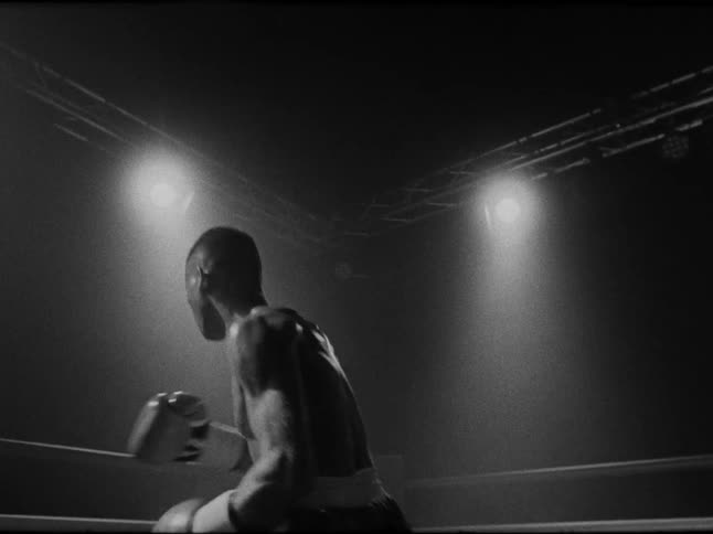 A shirtless male boxer stands in a ring under bright spotlights viewed from behind in black and white
