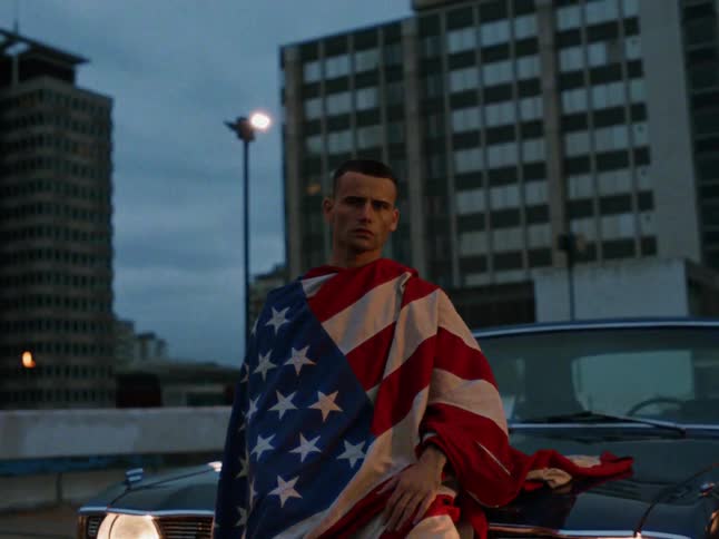 A man wrapped in an american flag leans against the hood of a classic blue car on a rooftop parking lot with tall city buildings and a street light in the background during twilight
