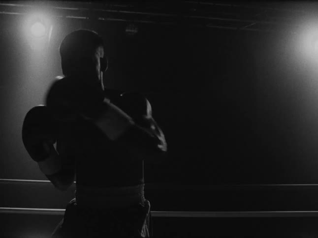 A man wearing boxing gloves moves around a dark boxing ring in a black and white scene with bright spotlights shining from above