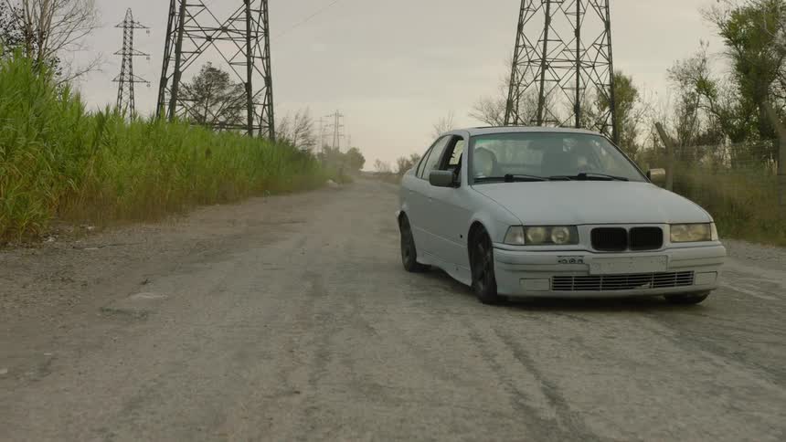 Two people wearing white faceless masks travel in a car on a dirt road between utility towers