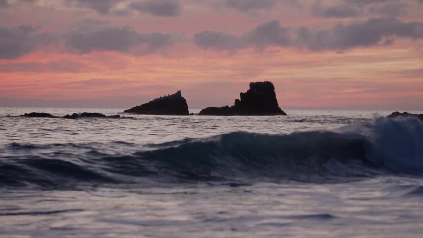 Dark rhythmic ocean waves roll and crash toward the shore in the foreground while jagged rock silhouettes stand firmly against a vibrant pink and orange sunset sky as peaceful twilight settles over the coast