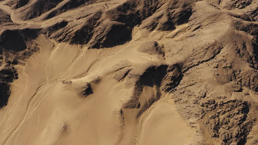 Drifting smoothly high above a vast and arid desert landscape the sweeping windblown sand dunes beautifully contrast with the deeply shadowed rugged rock formations bathed in warm golden sunlight
